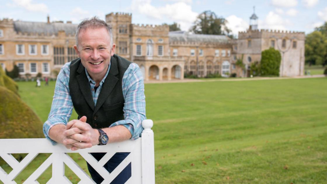 Man smiling at camera, leaning on a white fence with a large manor house in the background.